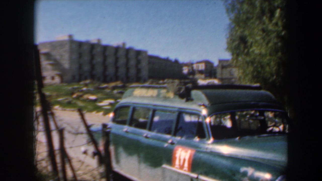 A green mid-century station wagon sits near a fence in Israel, with a red side marking and apartment blocks behind it.