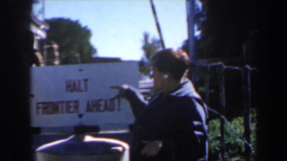 1960 Israel frame with a person pointing at a HALT FRONTIER AHEAD sign beside fencing and greenery.