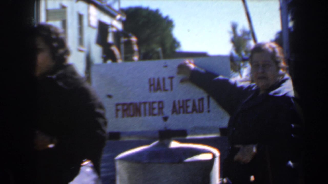 The woman faces the camera while pointing at the frontier warning sign.