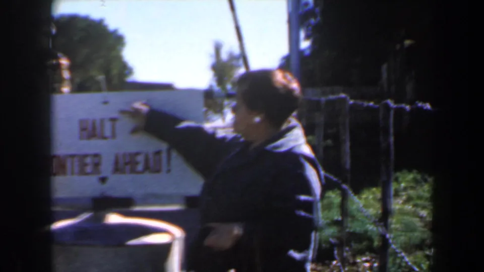 A side view shows the woman’s hand on the frontier warning sign while another figure passes close to the lens.