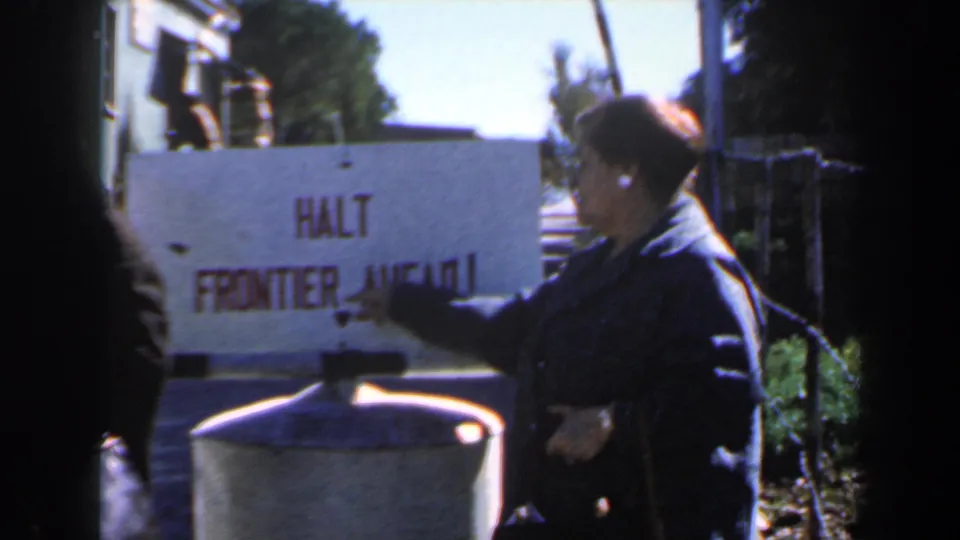 Near the end of the clip, the woman stands by the HALT FRONTIER AHEAD sign with trees and a street scene behind her.