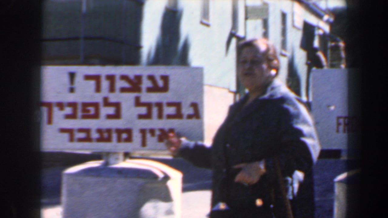 Woman in a dark coat points toward a bilingual frontier warning sign near concrete barriers.