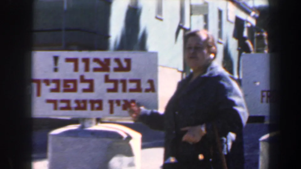 In 1960 Israel, a woman in a dark coat points at a Hebrew and English frontier warning sign beside concrete barriers.