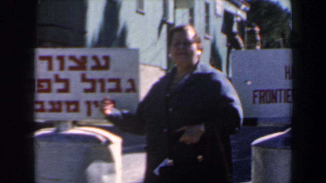 The woman remains beside the 1960 Israel frontier sign, with Hebrew lettering and part of the English warning visible.
