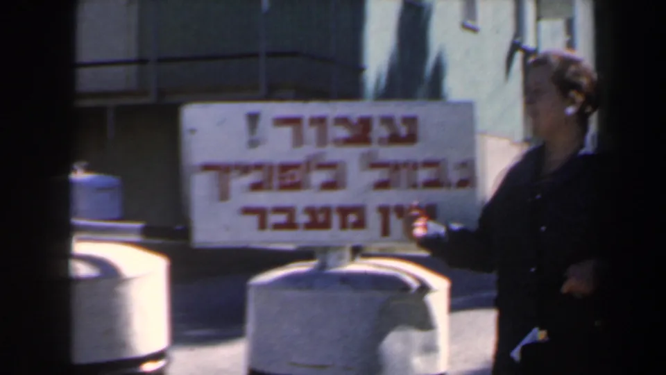 In the 1960 Israel home-movie frame, the woman gestures beside the Hebrew frontier sign and a concrete roadside barrier.