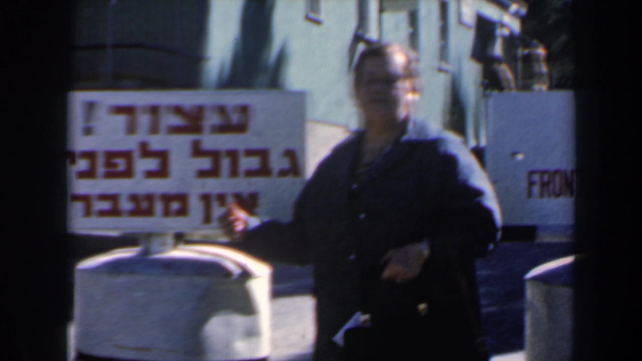 Woman holding a Hebrew sign near a frontier warning sign at a roadside checkpoint.