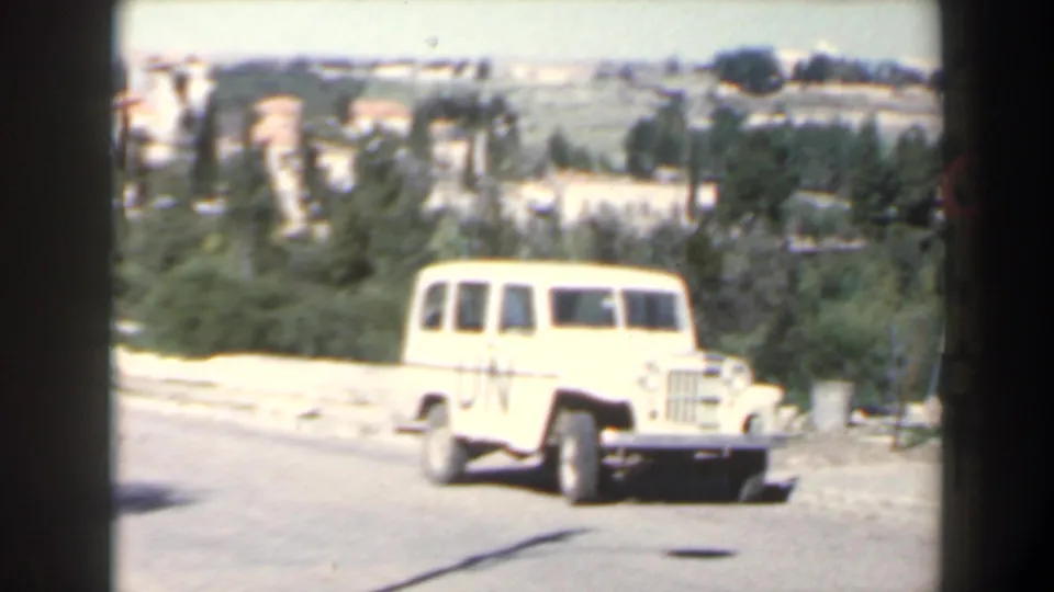 A white UN-marked jeep sits on a road in a hilly Israel landscape, seen in soft 8mm color footage.