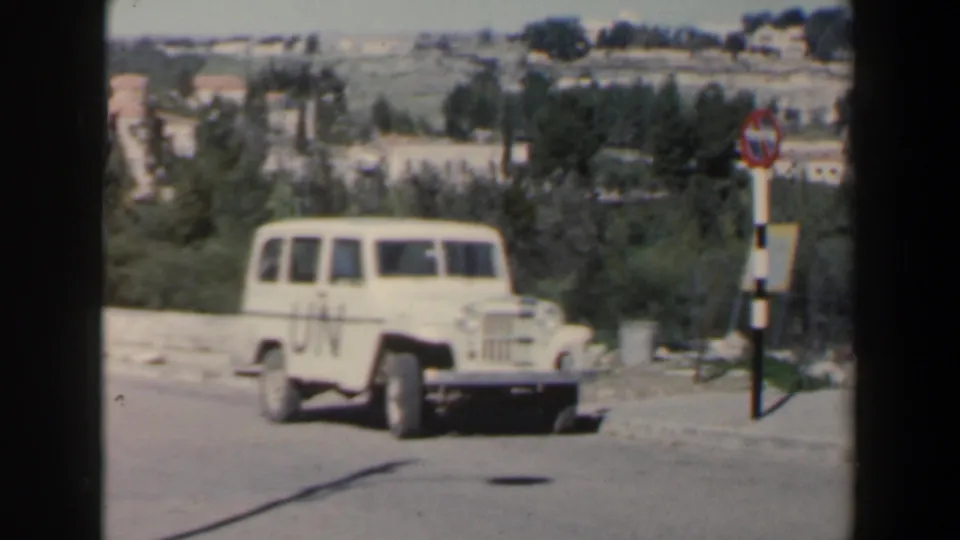 A white UN-marked jeep is framed beside a striped roadside sign, with hills and trees beyond in 1960s Israel footage.