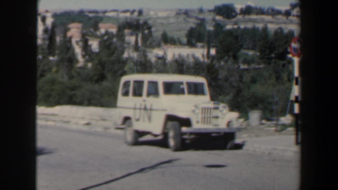 White UN-marked Jeep on a sunlit road with hillside buildings and trees behind it.