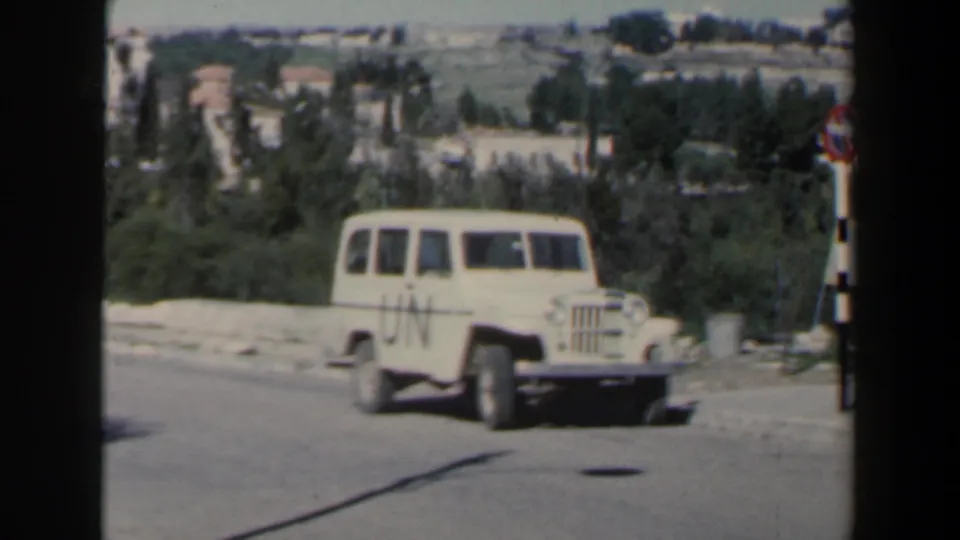 White UN-marked Jeep-style vehicle driving along a road in Israel, with trees, low buildings and distant hillside terrain visible.