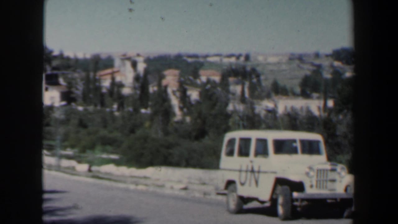 UN-marked Jeep near the right side of a road with hillside buildings and trees in the background.