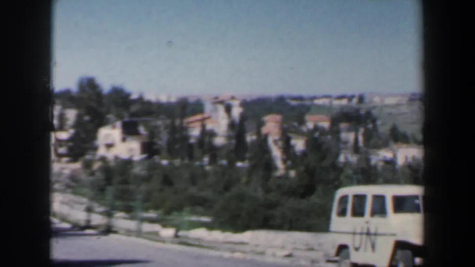 The white UN-marked Jeep appears at the lower right of the frame as the road, greenery and hillside buildings fill the background.