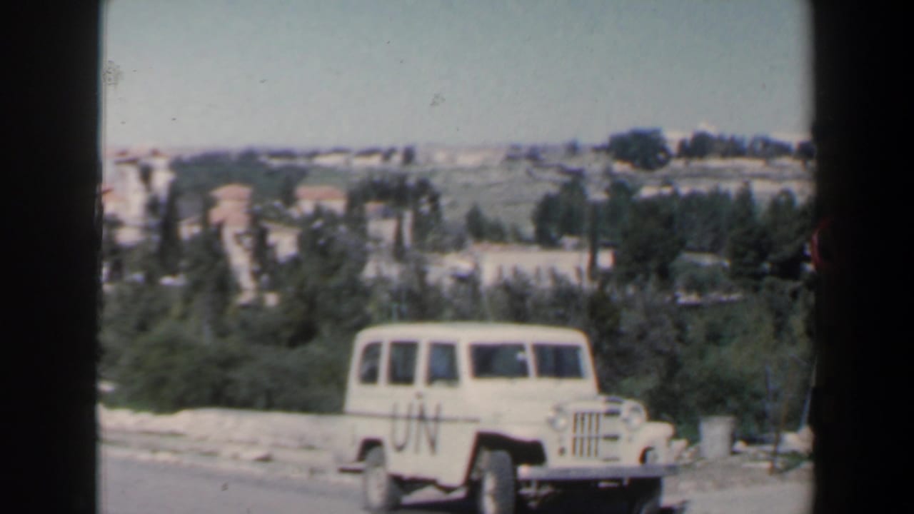 White UN-marked jeep on a sunlit road with trees and hills behind it.