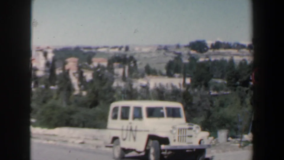 1960 Israel home-movie frame showing a white UN-marked jeep on a roadside against a hilly, tree-lined landscape.