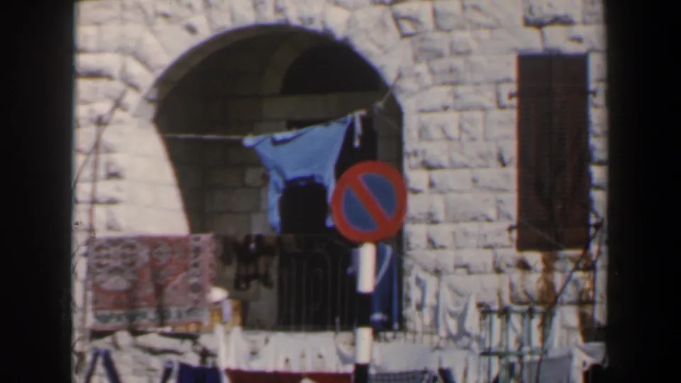 1960 Israel home-movie frame of an arched stone opening, laundry on a line, a rug or textile, and a red-blue no-parking sign.