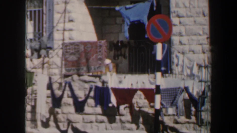 1960 Israel 8mm frame showing a stone facade, laundry on lines, barred windows, and a red-blue no-parking sign.