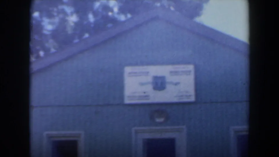 1960 Israel frame showing a green wooden building with a white sign, doorway openings, tree shade, and blue sky.
