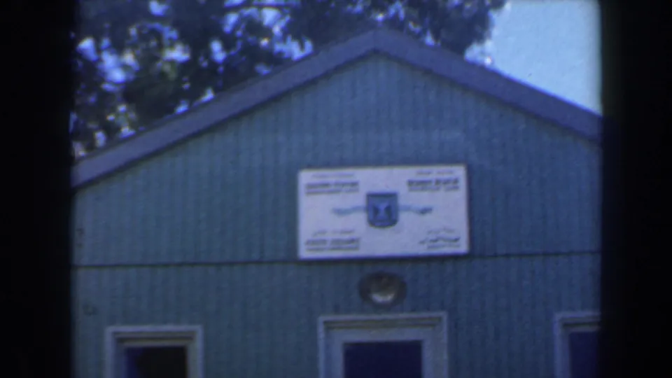 Clearer 1960 Israel 8mm view of a green facade with a white bilingual sign, open doorways, and trees overhead.