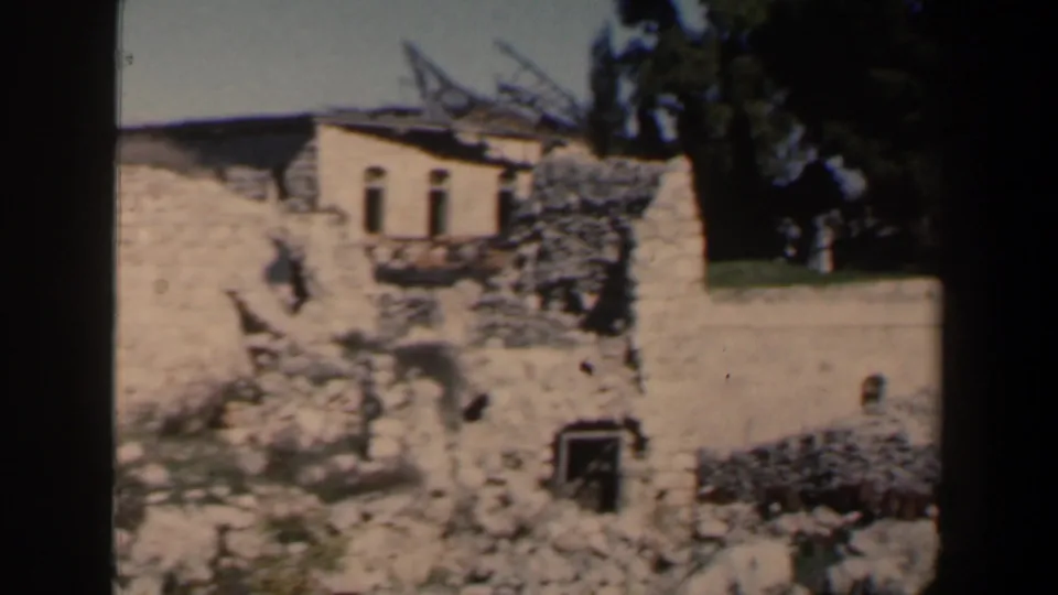 1960 Israel 8mm frame showing weathered stone ruins on a hillside, with a small pale structure and trees in bright daylight.