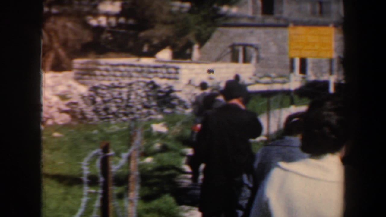 Visitors in period coats walk along a fenced path near stone walls, rubble, a building, and a yellow sign in Israel.
