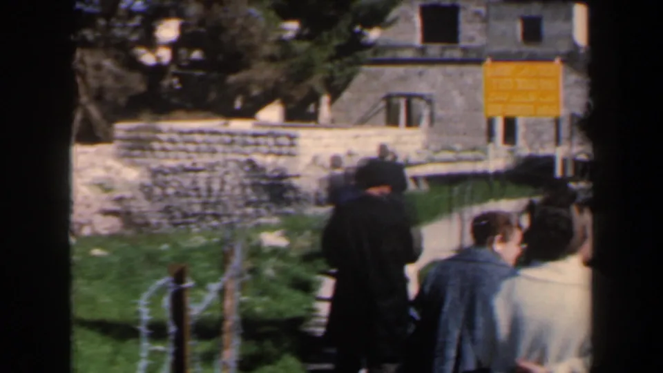A small group of visitors stands or walks near a wire fence, stone walls, grass, and a yellow sign at a historic-looking site in Israel.