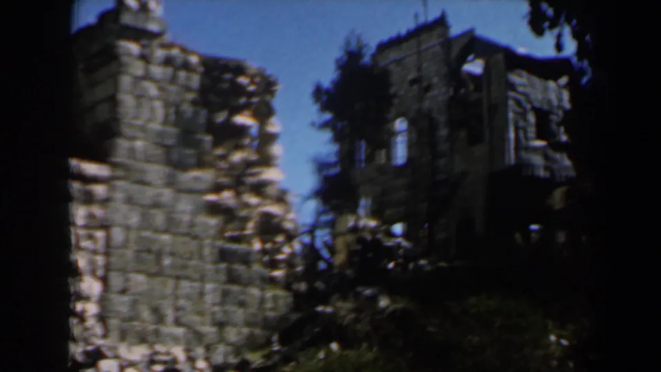 A tall weathered stone ruin stands against blue sky and trees, with deep shadow and handheld 8mm framing.