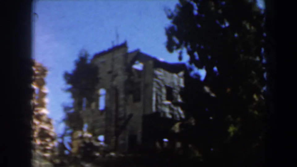 1960s Israel 8mm view of a grey stone ruin partly obscured by a dark tree trunk and foliage under blue sky.