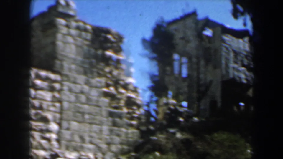 1960s Israel 8mm frame showing rough stone masonry and a damaged upper structure partly hidden by trees in daylight.