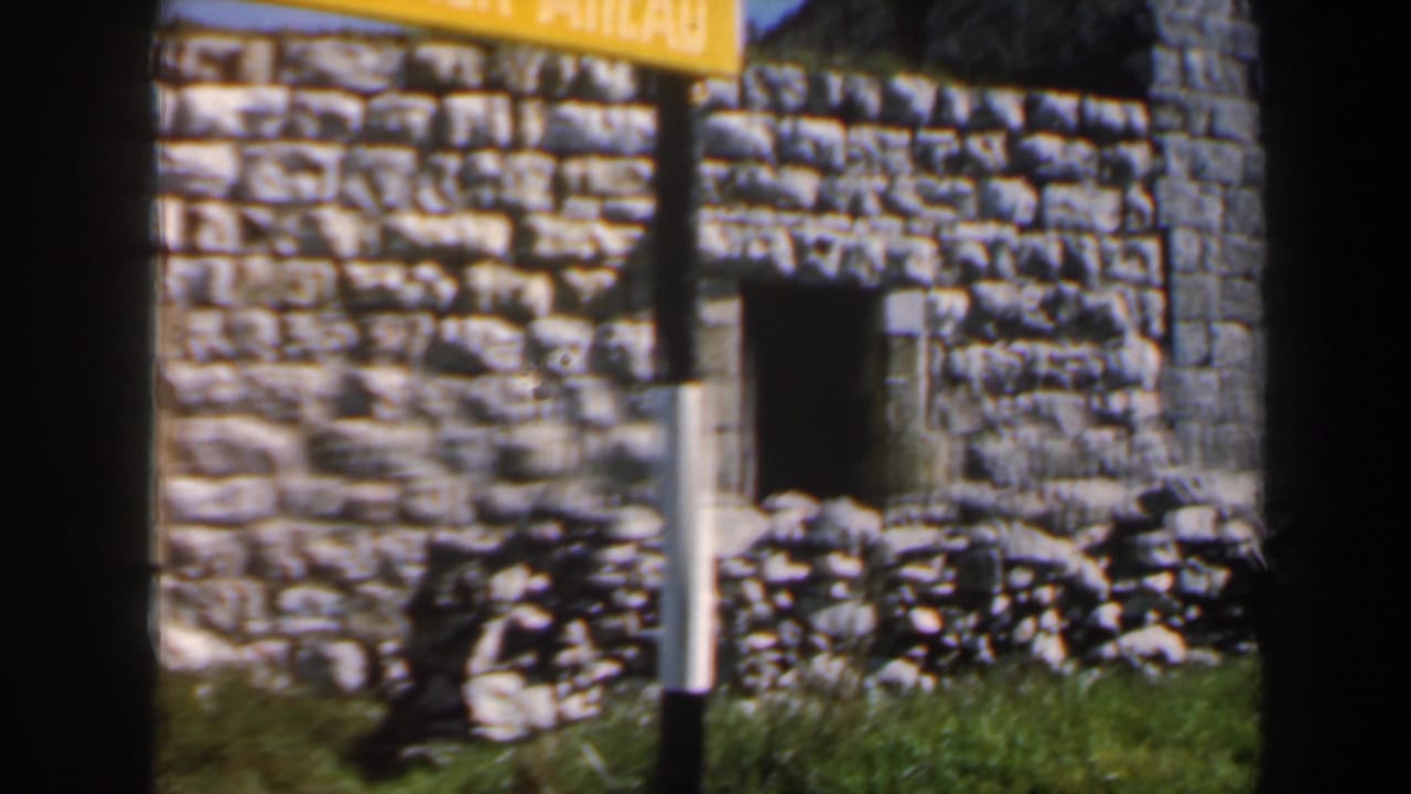 1960 Israel 8mm frame of a rough stone facade with a small opening, grass, and a partially visible yellow sign.