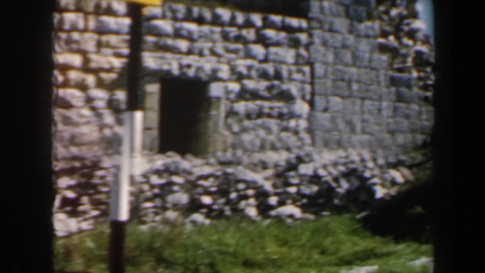1960 Israel 8mm frame showing a close view of rough stonework, a dark doorway, grass, rubble, and a black-and-white pole.