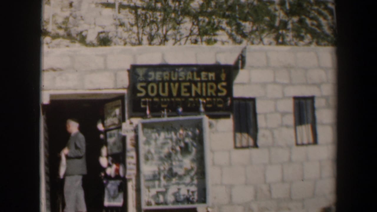 1960 Israel home-movie frame showing a suited man near a Jerusalem Souvenirs storefront with stone wall, doorway, and display board.