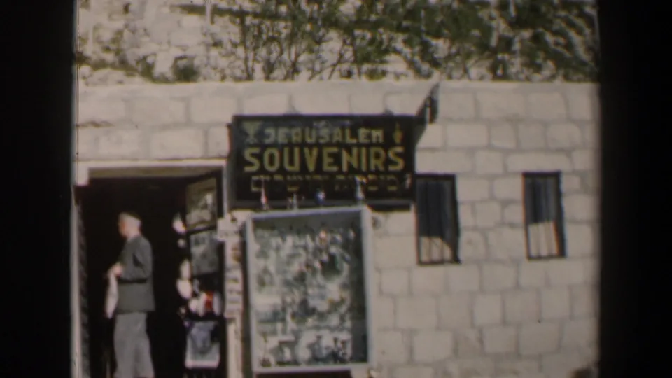 1960 Israel home-movie frame showing a suited man near a Jerusalem Souvenirs storefront with stone wall, doorway, and display board.