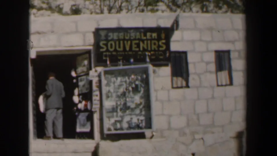 1960 Israel frame with a traveler standing at a stone souvenir shop doorway beneath English and Hebrew signage in Jerusalem.