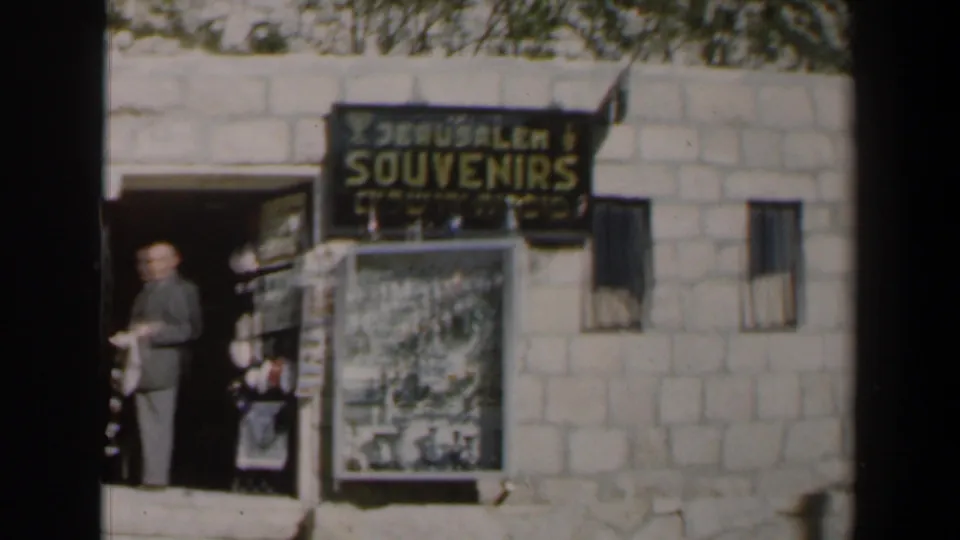 1960 Israel home-movie still of a Jerusalem souvenir storefront with a man at the doorway, large sign, display board, and window bars.