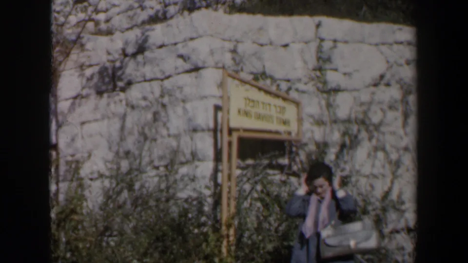 1960 Israel frame showing a traveler with a bag near rocky stonework and a sign for the King David Tomb in Jerusalem.