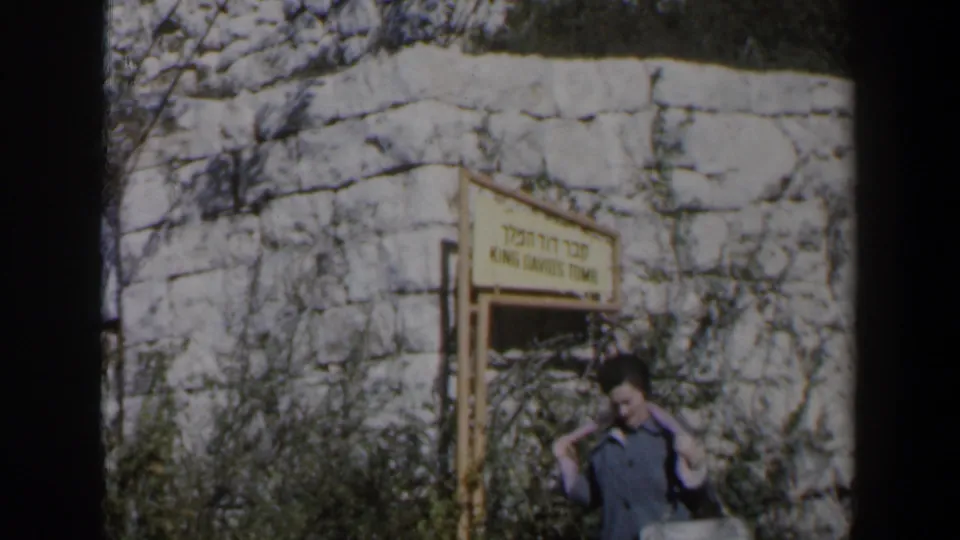 1960 Israel home-movie frame showing a woman near the King David's Tomb sign, with shrubs and a stone wall behind her.