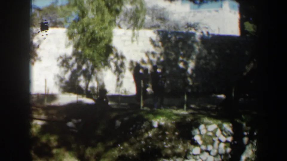 1960 Israel home-movie frame showing silhouetted figures by a bright white wall, trees, and a stone-edged garden area.