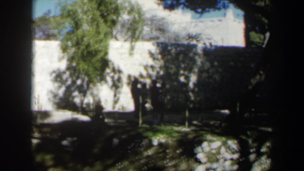 1960 Israel home-movie view of people in shadow near a white wall, with trees and garden stones in the foreground.