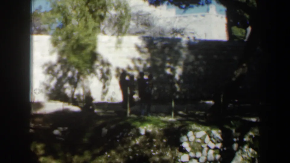 1960 Israel 8mm frame of silhouetted people near a white wall, framed by trees, garden planting, and strong sunlight.