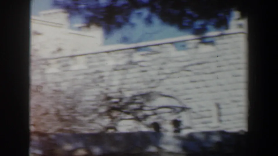 1960 Israel home-movie frame with motion blur on a pale exterior wall, tree shadows, and two small figures near the bottom edge.