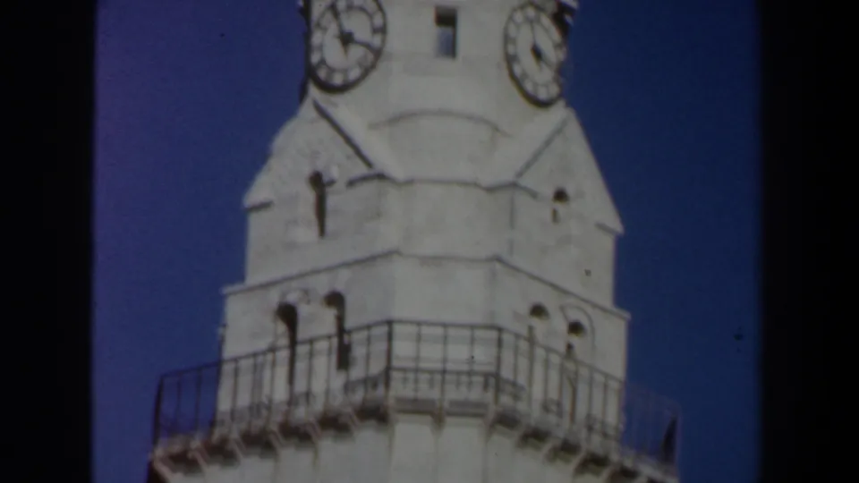 1960s Israel home-movie view of a white church tower top with clock faces and railing against deep blue sky.