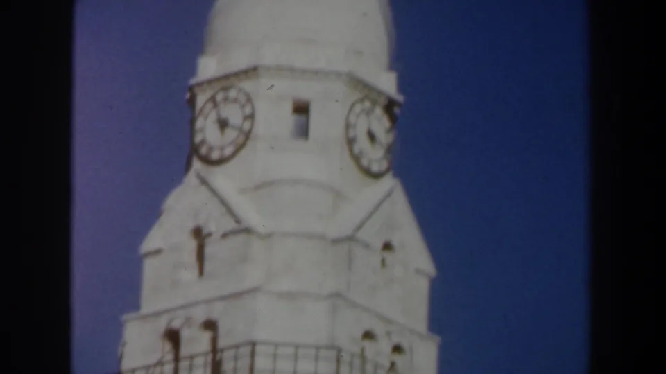 1960 Israel 8mm frame showing the upper portion of a white stone clock tower with two clock faces against clear blue sky.