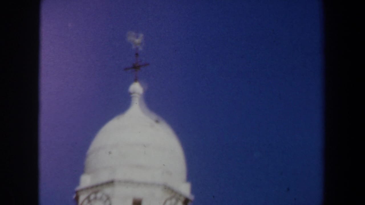 1960 Israel 8mm frame showing the domed top of a pale stone tower, balcony railing, and weather vane against blue sky.
