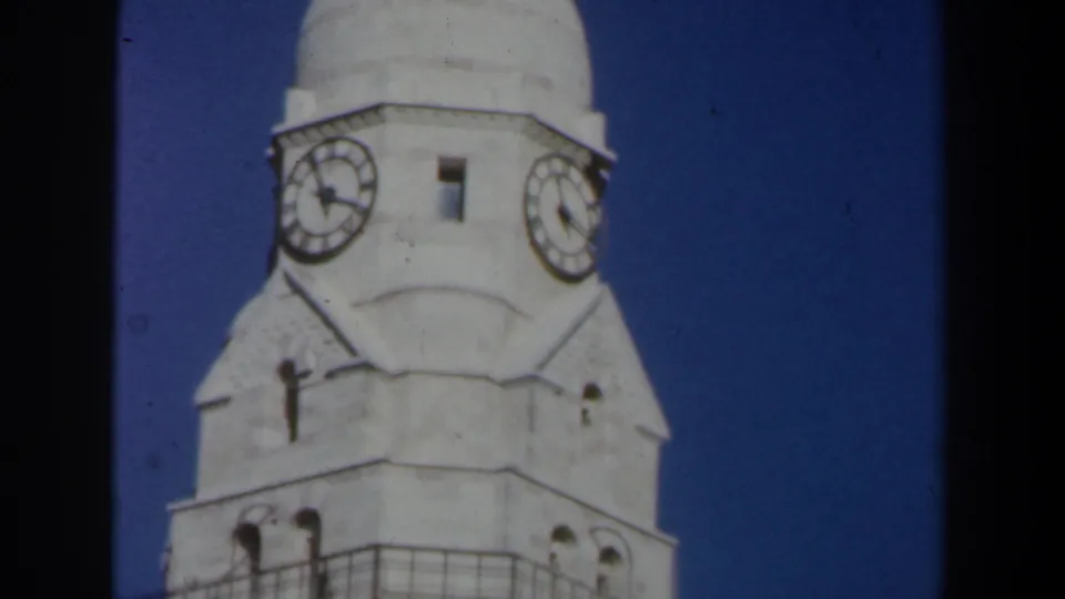 1960 Israel 8mm frame with a closer view of the clock tower's clock faces, pale masonry, ledges, and railings.