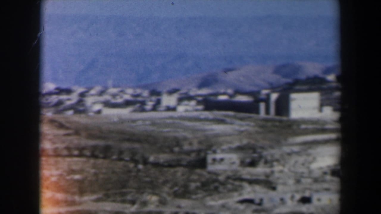 Distant white buildings across arid hills under a blue sky, framed by dark film edges.