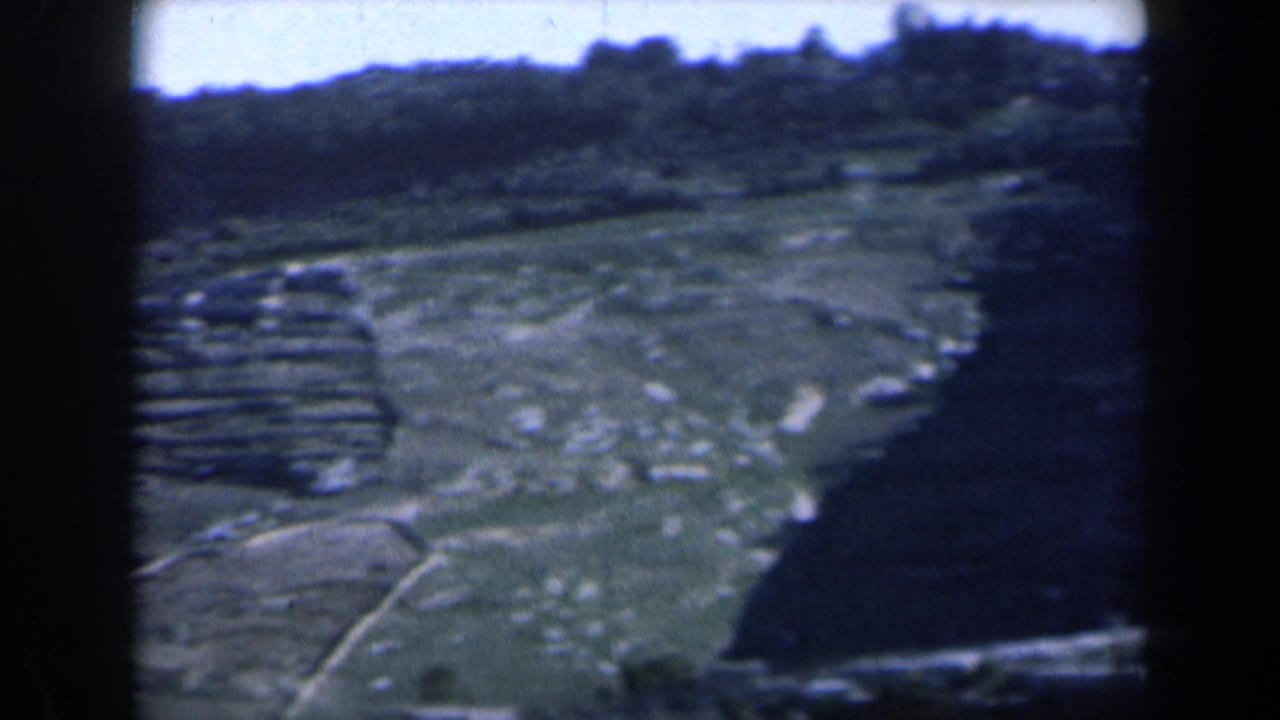 Hilly landscape with terraced slopes, scattered rocks, vegetation, and a dark structure edge.