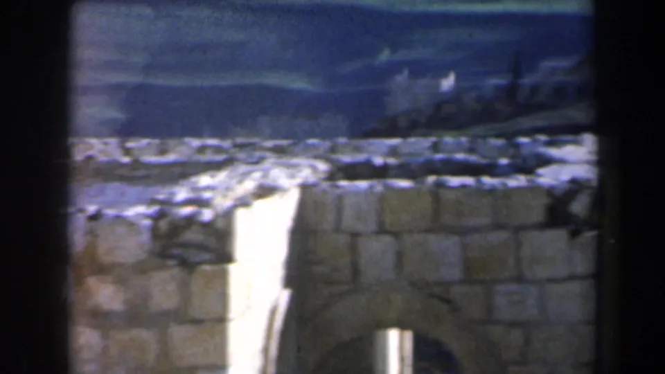 1960 8mm frame in Israel showing a stone wall or ruin in the foreground with distant hillside structures and open terrain behind it.
