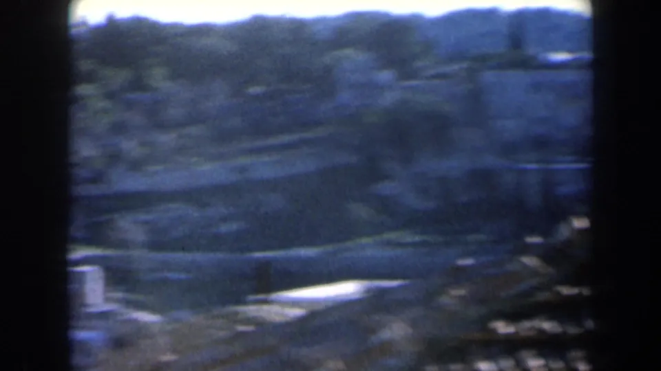 1960 Israel home-movie frame showing a soft, blue-tinted panorama of hilly terrain with paths, vegetation, and low structures.