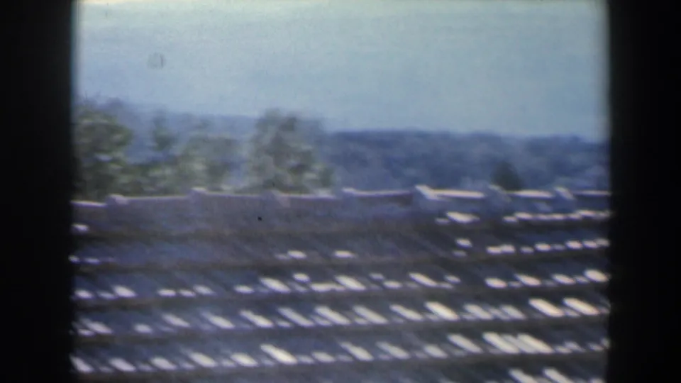 1960 Israel home-movie frame with a hazy rooftop foreground, pale wall, scattered trees, and distant terrain.