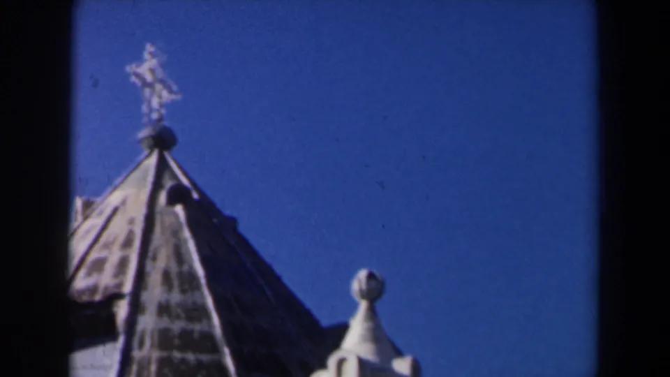 1960 Israel 8mm frame showing a roof peak topped by a cross, with a pale turret and blue sky in the background.